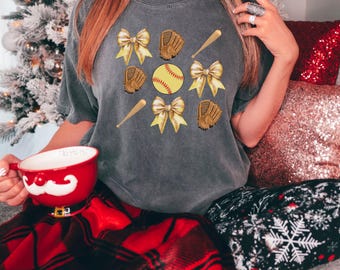 May include: A gray t-shirt with a softball design. The design features a softball, bats, gloves, and bows. The bows are gold and the softball is yellow with red stitching.