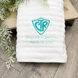 May include: White bath towel with a teal embroidered shield with the letters CTR inside. The towel is personalized with the name Brynlee R Barlon and the date March 18, 2023.