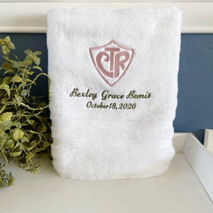 May include: White bath towel with a pink embroidered shield with the letters "CTR" inside. The text "Bexley Grace Remis October 18, 2020" is embroidered below the shield.