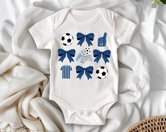 May include: White baby bodysuit with blue soccer-themed graphics. The design includes soccer balls, bows, a jersey, a foam finger with the number one, and the word "GOAL" with a soccer net.