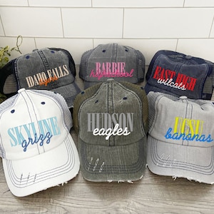 May include: A collection of six distressed trucker hats with different embroidered designs. The hats are in various colors, including white, gray, and blue. The designs include "Idaho Falls Tigers", "Barbie High School", "East High Wildcats", "Skyline Grizz", "Hudson Eagles", and "UCST Bananas".