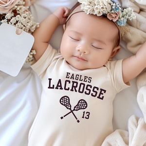 May include: A sleeping baby wearing a beige onesie with the words "EAGLES LACROSSE" in dark purple. The onesie features crossed lacrosse sticks and the number 13. The baby is wearing a floral headband and is lying on a white surface.