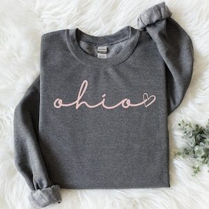 May include: A dark gray sweatshirt with the word "ohio" in a cursive, light pink font. A small pink heart is next to the "o". The sweatshirt is folded on a white, fluffy surface.