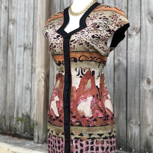 May include: A patterned dress with a V-neckline and short sleeves. The dress features a button-down front with black trim and a gold-colored necklace. The fabric has a mix of colors, including brown, beige, and pink, with a floral and geometric design.