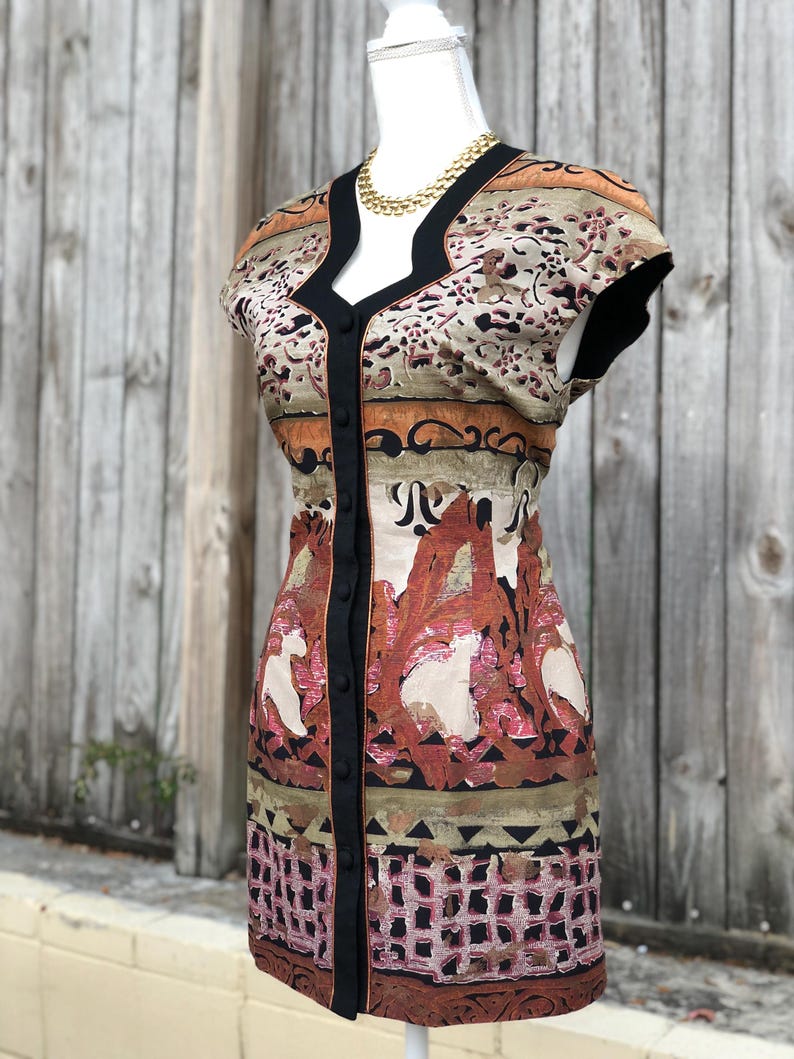 May include: A patterned dress with a V-neckline and short sleeves. The dress features a button-down front with black trim and a gold-colored necklace. The fabric has a mix of colors, including brown, beige, and pink, with a floral and geometric design.