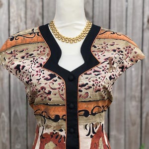 May include: A vintage-style top with a unique pattern in shades of beige, orange, and burgundy, accented with black trim and buttons. A gold-toned necklace complements the neckline. The top is displayed on a mannequin.