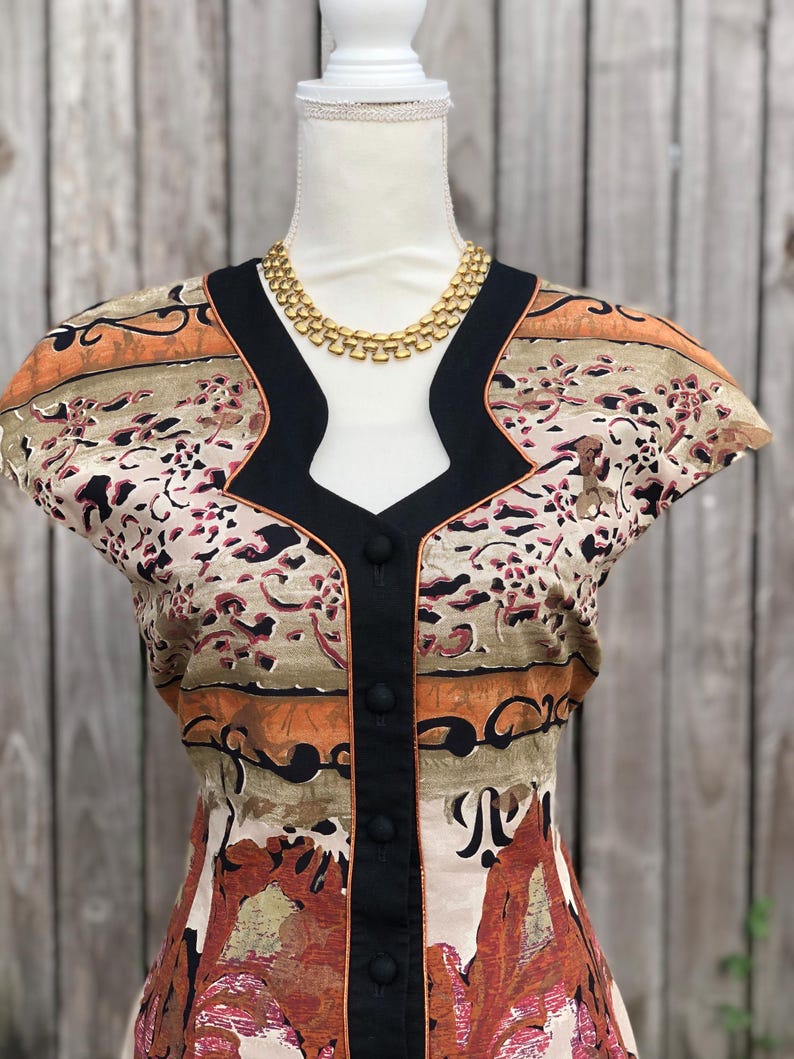 May include: A vintage-style top with a unique pattern in shades of beige, orange, and burgundy, accented with black trim and buttons. A gold-toned necklace complements the neckline. The top is displayed on a mannequin.
