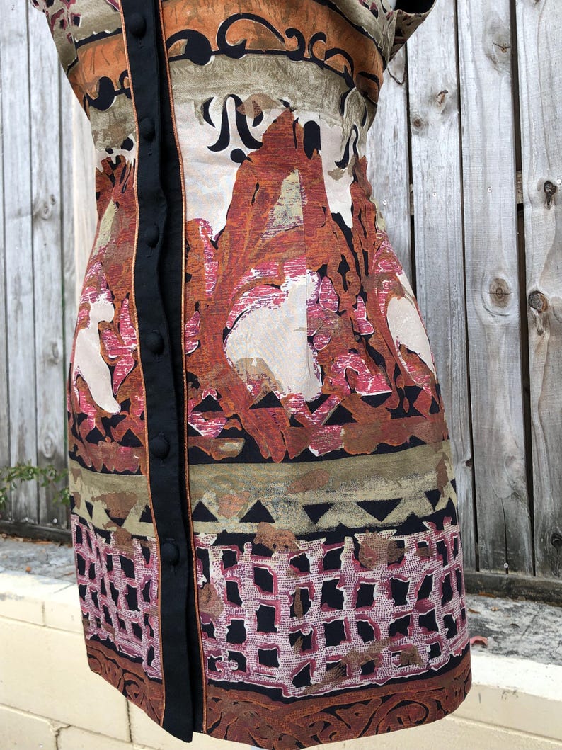 May include: A patterned dress with a button-up front. The dress features a mix of colors, including orange, brown, and black, with a geometric and floral design. The dress has a black trim and buttons.