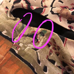 May include: Close-up of a section of fabric with a complex pattern. The design features a mix of colors, including tan, black, red, and orange. The fabric appears to be folded, revealing a black lining. The pattern includes abstract shapes and animal-like figures.