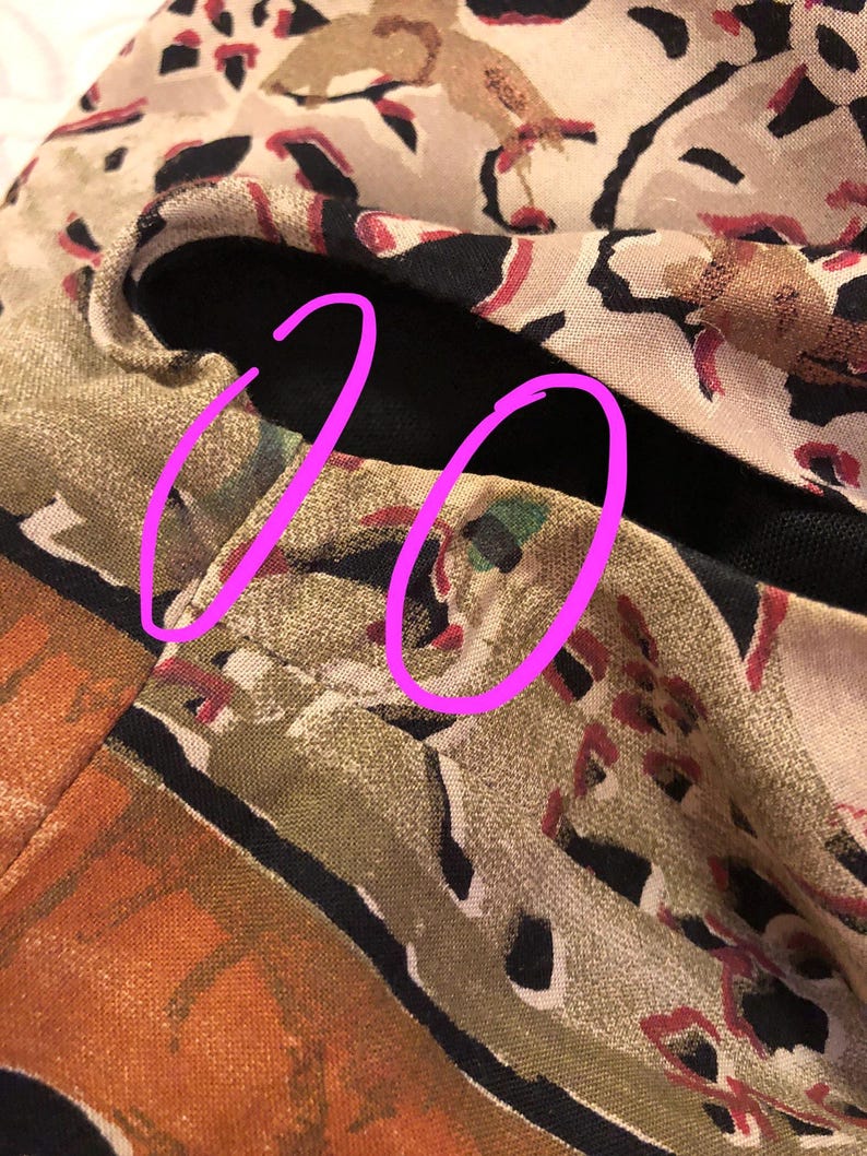 May include: Close-up of a section of fabric with a complex pattern. The design features a mix of colors, including tan, black, red, and orange. The fabric appears to be folded, revealing a black lining. The pattern includes abstract shapes and animal-like figures.