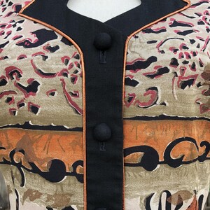 May include: Close-up of a vintage patterned jacket. The jacket features a beige, brown, and red abstract design. It has a black collar and button closure, with a thin orange trim. The jacket is a unique piece of vintage apparel.