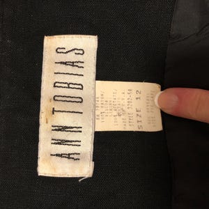 May include: Close-up of a black garment with two attached tags. The larger tag reads "ANN TOBIAS" in white capital letters. The smaller tag provides fabric content, size 12, and style information. The garment's fabric is black.