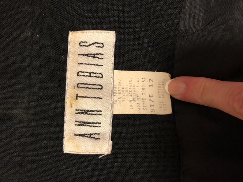 May include: Close-up of a black garment with two attached tags. The larger tag reads "ANN TOBIAS" in white capital letters. The smaller tag provides fabric content, size 12, and style information. The garment's fabric is black.