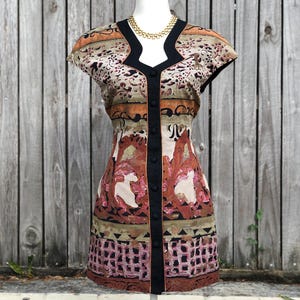 May include: A patterned mini dress with a black trim and button closure. The dress features a V-neckline and cap sleeves. The fabric has a mix of colors, including brown, beige, and pink, with a geometric and floral design. A gold necklace is visible.