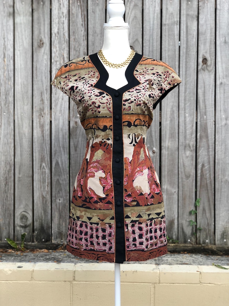 May include: A patterned mini dress with a black trim and button closure. The dress features a V-neckline and cap sleeves. The fabric has a mix of colors, including brown, beige, and pink, with a geometric and floral design. A gold necklace is visible.
