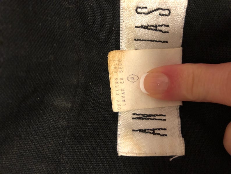 May include: Close-up of a black fabric with two white labels. One label reads "SAYI" in black capital letters. The other label is a care instruction tag with the words "DRY CLEAN ONLY" and "LAVAR EN SECO".