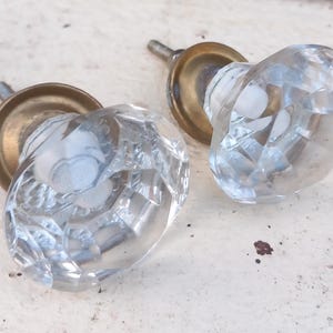 Puede incluir: Dos pomos de puerta de cristal transparente y facetado con bases color latón. Los pomos tienen un patrón de diamantes y están montados sobre una superficie blanca. Las bases de latón muestran signos de edad.