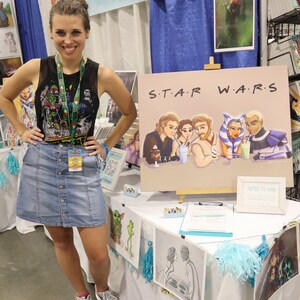 Peut inclure: Une femme se tient devant une table pr&eacute;sentant des &oelig;uvres d'art. La table est recouverte d'une nappe blanche avec des bordures bleues et jaunes. Les &oelig;uvres d'art comprennent une pi&egrave;ce sur le th&egrave;me de Star Wars avec le texte "S.T.A.R. W.A.R.S." et une pancarte indiquant "Entrez pour gagner".
