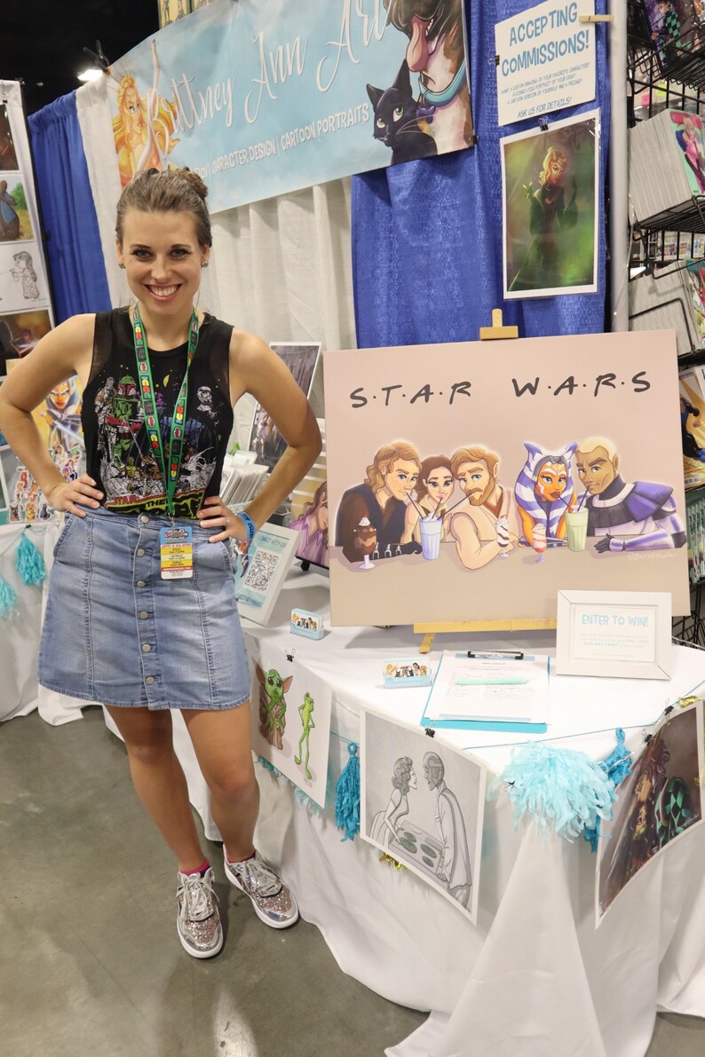 Peut inclure: Une femme se tient devant une table pr&eacute;sentant des &oelig;uvres d'art. La table est recouverte d'une nappe blanche avec des bordures bleues et jaunes. Les &oelig;uvres d'art comprennent une pi&egrave;ce sur le th&egrave;me de Star Wars avec le texte "S.T.A.R. W.A.R.S." et une pancarte indiquant "Entrez pour gagner".