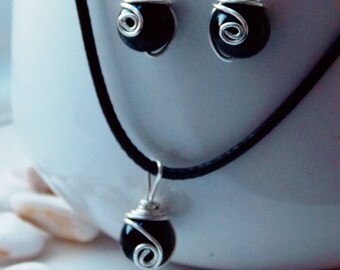 May include: A jewelry set featuring a necklace and matching earrings. Each piece has a black, spherical bead wrapped in silver-colored wire. The necklace hangs on a black cord, and the earrings have silver hooks.
