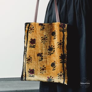 May include: A rectangular tote bag made of mustard yellow velvet fabric featuring a repeating pattern of small dark floral motifs. The bag includes two thin brown leather shoulder straps and black vertical side panels along the edges.