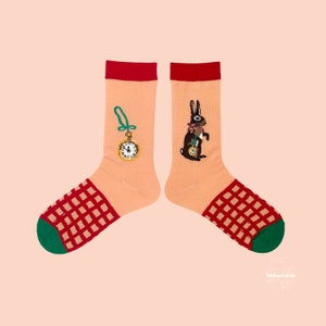 May include: A pair of peach-colored socks with red and green accents. One sock features a gold pocket watch, the other a black rabbit holding a pocket watch. The toe area has a red and green checkered pattern. The socks are a whimsical accessory.