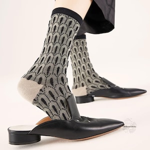 May include: Black leather mules with a pointed toe and low heel. The feet are wearing patterned socks in grey and black. The socks feature a repeating geometric design. The shoes are a size 38.