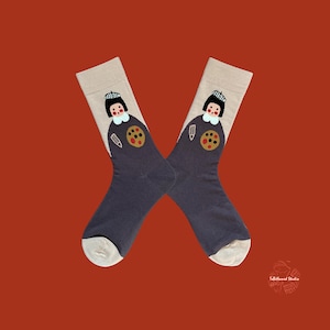 May include: A pair of navy blue socks with a beige cuff and toe. Each sock features a cartoon illustration of a person with a black hat, white collar, and a gold circle with a brown design. A pencil is also included in the design.