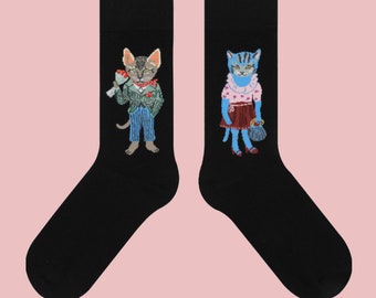 Op de afbeelding: Een paar zwarte sokken met geborduurde kattenontwerpen. Eén sok toont een kat in een pak met bloemen, de andere een kat in een jurk met een handtas. De achtergrond is lichtroze.