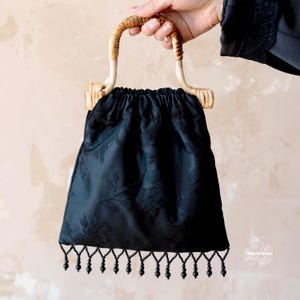 May include: A black drawstring handbag featuring a tonal floral jacquard pattern, a curved light brown rattan handle, and a bottom edge finished with a row of dangling black beads. The bag has a gathered top closure and a structured silhouette.