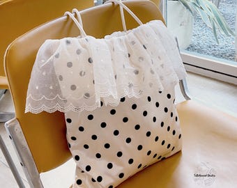 Puede incluir: Un bolso tote de color crema con un patrón de lunares negros y una capa de tul blanco transparente con escote de volantes y bordes festoneados. El bolso tiene dos correas finas y el texto ToBeNamed Studio.