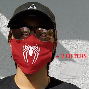 May include: Red face mask with a white spider logo and a honeycomb pattern. Includes two PM2.5 filters. The text "+ 2 FILTERS" is visible in red. The person is wearing a black cap and sunglasses.
