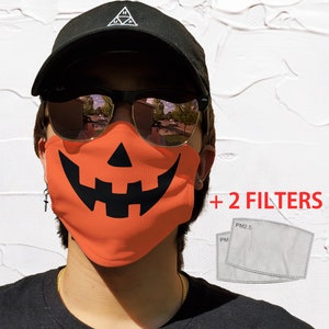 May include: Orange face mask with a black jack-o'-lantern design. The mask comes with two white PM2.5 filters. + 2 FILTERS is written in red text.
