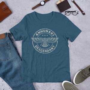 Pu&ograve; includere: Una t-shirt blu turchese con una grafica bianca invecchiata di un simbolo alato con il testo "ANNUNAKI DESCENDANT" in un cerchio.