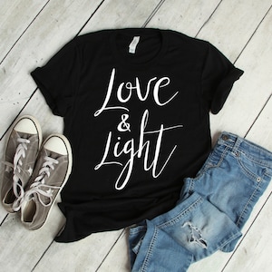 Könnte beinhalten: Schwarzes T-Shirt mit weißem Text "Love & Light" auf weißem Holzgrund. Das Shirt liegt auf einem Paar grauen Sneakern und einer blauen Jeans.