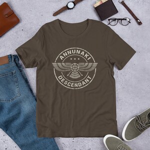 Pu&ograve; includere: Una t-shirt marrone con una grafica bianca invecchiata di un cerchio alato con il testo "ANNUNAKI DESCENDANT" all'interno.
