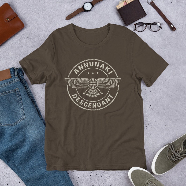 Pu&ograve; includere: Una t-shirt marrone con una grafica bianca invecchiata di un cerchio alato con il testo "ANNUNAKI DESCENDANT" all'interno.