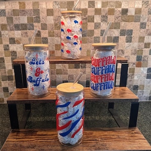 May include: Four clear glass tumblers with bamboo lids and glass straws. One features red and blue buffalo silhouettes, one says Let's Go Buffalo with stars, one repeats the word BUFFALO in red and blue, and one has red and blue diagonal stripes.