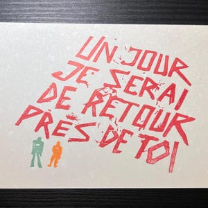 Pode incluir: Um cartão branco com texto vermelho que diz "UN JOUR JE SERAI DE RETOUR PRES DE TOI". Duas pequenas figuras são verdes e laranja. O cartão está sobre uma superfície escura.