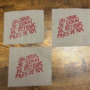 Könnte beinhalten: Drei quadratische, hellgraue Stoffflicken mit rotem Text im handschriftlichen Stil: "UN JOUR JE SERAI DE RETOUR PRES DE TOI". Die Flicken haben eine strukturierte Oberfläche und einen dekorativen Zickzackrand.