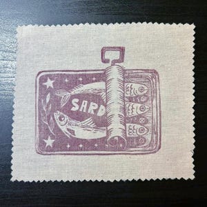 May include: A square, off-white fabric patch featuring a purple print of a sardine can. The can is open, showing fish. The word "SARD" is printed above the fish. Stars and leaves surround the can.