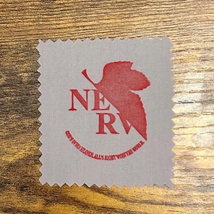 May include: A square gray microfiber cleaning cloth with pinked edges. The center features a red graphic of a leaf shape behind the text NERV and the circular text God's in his heaven, all's right with the world printed in red ink.