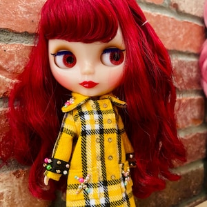 May include: A doll with long, wavy, vibrant red hair and large eyes. The doll wears a yellow, black, and white plaid coat featuring a double-breasted design with metallic buttons and small, colorful beaded accents on the cuffs and pockets.