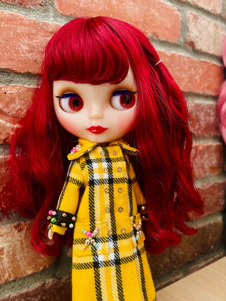 May include: A doll with long, wavy, vibrant red hair and large eyes. The doll wears a yellow, black, and white plaid coat featuring a double-breasted design with metallic buttons and small, colorful beaded accents on the cuffs and pockets.