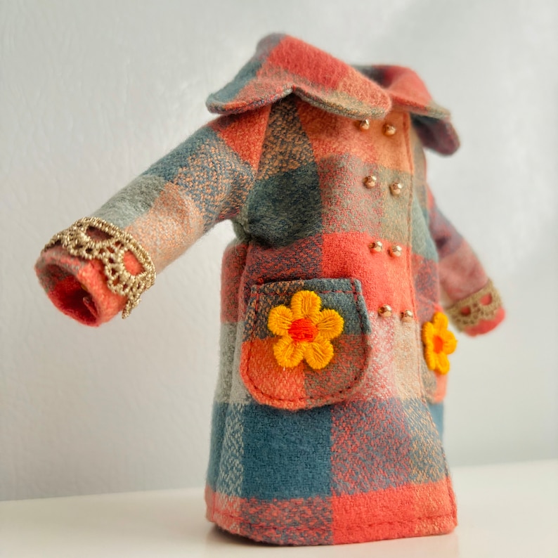 May include: A red and blue plaid coat with a large collar and two yellow flower embellishments. The coat has gold buttons and a lace trim on the sleeves.