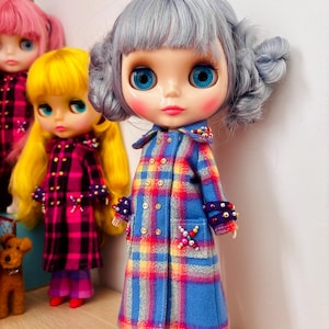 以下が含まれることがあります： グレーの髪の毛をした人形が、青、黄、赤のチェック柄のコートとそれに合うズボンを着ています。コートには装飾的なボタンと装飾が施されています。ピンクと黄色の髪の毛をした他の人形が背景にあります。