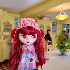 May include: A red-haired doll with a pink and white patterned dress and a pink and white floral hat. The doll has large blue eyes and is standing in a room with yellow walls and a white table.