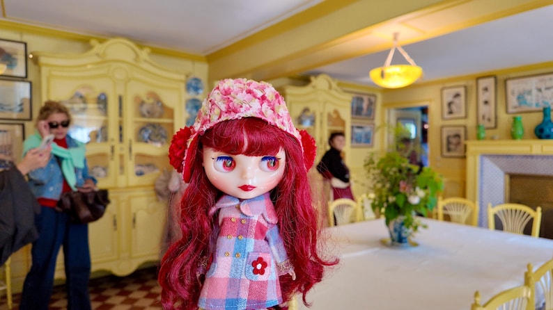 May include: A red-haired doll with a pink and white patterned dress and a pink and white floral hat. The doll has large blue eyes and is standing in a room with yellow walls and a white table.