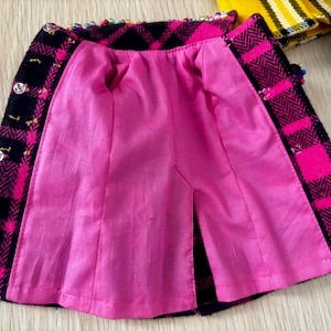 May include: A miniature skirt featuring a black and hot pink plaid pattern with a herringbone texture. The garment is lined with bright pink fabric and includes small silver-toned buttons along the front edge and colorful beads along the waistband.