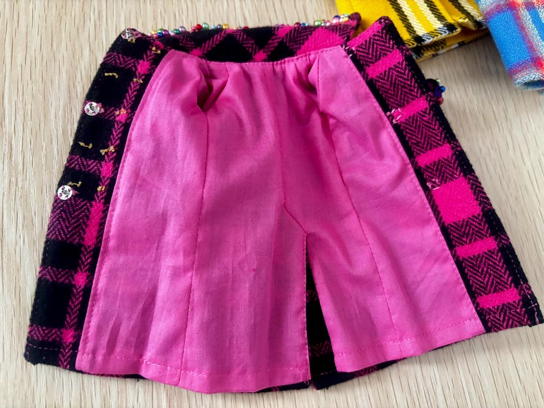 May include: A miniature skirt featuring a black and hot pink plaid pattern with a herringbone texture. The garment is lined with bright pink fabric and includes small silver-toned buttons along the front edge and colorful beads along the waistband.